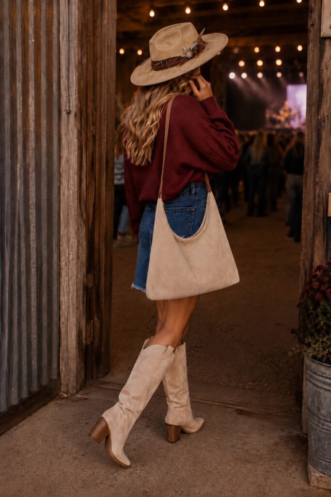 Woman wearing a burgundy sweater with textured sleeves, button-front denim skirt, tan western hat, cream suede boots, slouchy neutral shoulder bag, and gold jewelry styled as an elevated western outfit for country concert nights and fall festivals.